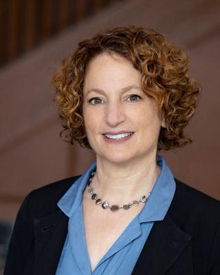 Headshot of a white woman with curly hair. She is smiling and wearing a blue button-down and black blazer.