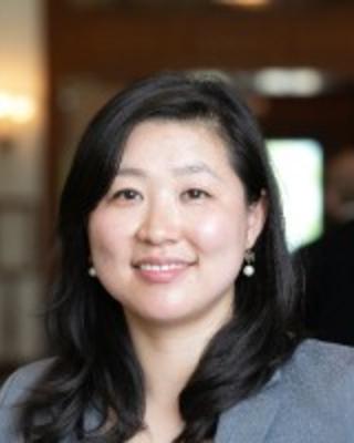 A professional headshot of an Asian woman with dark hair that falls past her shoulders and light skin. She is smiling and wearing a gray blazer.